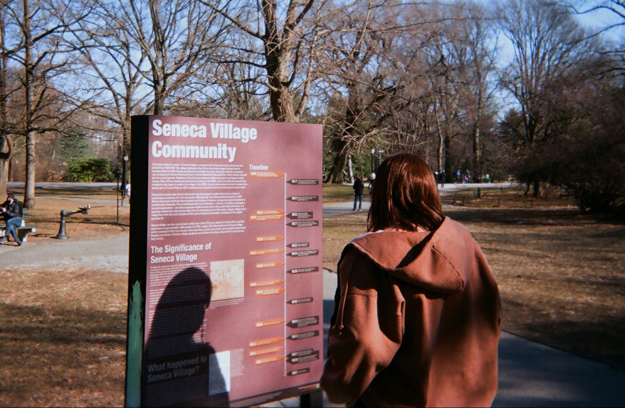 A figure reading the Seneca Village Community historical marker in Central Park, winter trees behind her